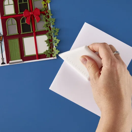 Hand hält das weiße Spellbinders Comfort Craft Creasing Tool zum Falzen von weißem Papier neben einer gebastelten Karte.