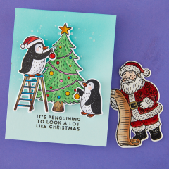 Türkisfarbene Karte mit zwei kolorierten Pinguinen, die einen Christbaum mit Kugeln schmücken, und Grußtext 'It's penguining to look a lot like Christmas'.