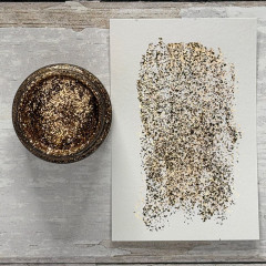 Offene Dose Tim Holtz Distress Liquid Tinsel Antiqued neben aufgestrichenem Muster auf Papier, zeigt feinen Gold-Silber-Glitter und Textur