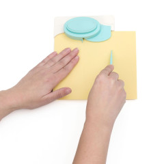 Hände verwenden das Rillwerkzeug auf gelbem Papier mit dem hellblauen Pillow Box Punch Board oben