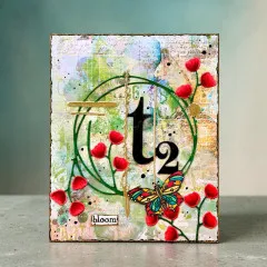 Grün-rote Karte mit Mohnblüten, buntem Schmetterling und der kreisförmigen „t2"-Stanzform mit Knospenzweig, Wortsticker „bloom"