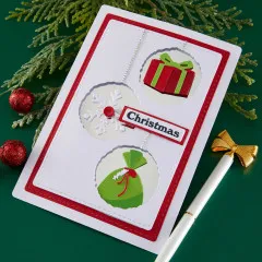 Weihnachtskarte mit drei hängenden Kugeln aus gestanztem Geschenk, Schneeflocke und Sack sowie dem Schriftzug 'Christmas'.