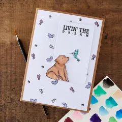 Grußkarte mit Bärenjunges und Kolibri umgeben von Schmetterlingen, Spruch 'Livin' The Dream'.