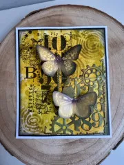 Gelbe Mixed-Media-Karte mit zwei Schmetterlingen, Zahnrad-Stempeln und Schriftzug „Wildlife" auf Holzscheiben-Untergrund