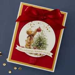 Anwendungsbeispiel: Weihnachtskarte mit Teddybär-Rub-On, roter Satinschleife und Prägeschriftzug 'Delivering Warm Hugs'.