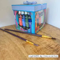 Blaue Geschenkbox mit rundem Fenster, gefüllt mit Acrylfarbtuben, daneben zwei Pinsel