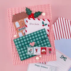 Detailaufnahme der weihnachtlichen Postkarte mit Lebkuchenhaus, 'Dear Santa'-Brief und kariertem Umschlag auf geprägtem Hintergrund.
