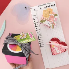 Rosa Geschenkbox und Grußkarte mit Herzen, Falzbrett und Falzbein neben Dekoration
