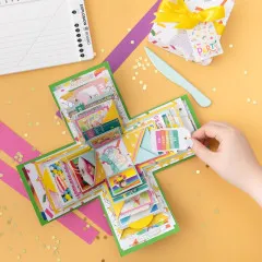Bunte Explosionsbox aus grünem und gelbem Papier mit aufgeklappten Fächern, Hand deutet auf Inhalte