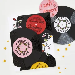 Nahaufnahme einer Weihnachtskarte aus schwarzen Glitzer-Schallplatten mit farbigen Labeln 'A Merry Little Christmas' und 'A Holly Jolly Christmas' sowie Lichterketten-Stanzteilen.