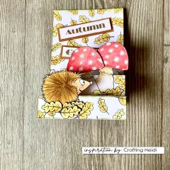 Stufenkarte „Autumn" mit Igel zwischen zwei roten Fliegenpilzen vor gelbem Herbstblätter-Muster.