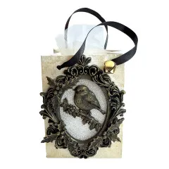 Helle Geschenktüte mit angehängtem bronzefarbenem Ovalrahmen, Glitzerhintergrund und Rotkehlchen-Figur sowie Glöckchen am Band
