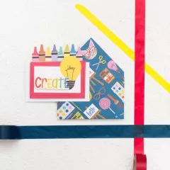Bunte Mini-Umschläge mit 'Create' Rahmen und Glühbirnen-Motiv, Stifte und Bastelmaterial im Hintergrund