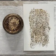 Offene Dose Tim Holtz Distress Liquid Tinsel Antiqued neben aufgestrichenem Muster auf Papier, zeigt feinen Gold-Silber-Glitter und Textur