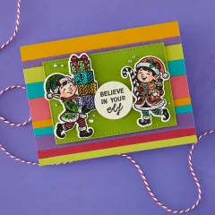 Bunt gestreifte Karte mit zwei kolorierten Elfen - eine trägt Geschenke, die andere ein Kuscheltier - grünem Rahmen und Spruch 'Believe in your elf'.