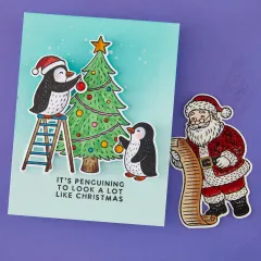 Türkisfarbene Karte mit zwei kolorierten Pinguinen, die einen Christbaum mit Kugeln schmücken, und Grußtext 'It's penguining to look a lot like Christmas'.