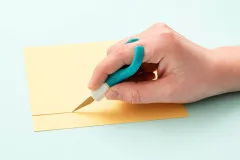 Hand schneidet gelbes Bastelpapier mit Comfort Craft Messer, ergonomischer Fingergriff deutlich sichtbar