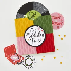 Colorblock-Karte in Gelb, Grün, Rosa und Rot mit glitzernder Schallplatte in grüner Hülle, kleinem Retro-Plattenspieler und Grußtext 'Cue the Holiday Tunes'.