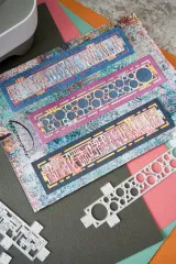 Mixed-Media-Karte mit drei geschichteten Sockets-Bordüren in Blau, Pink und Gold, daneben die Stanzen