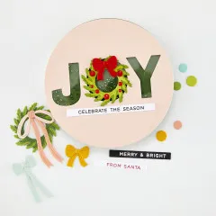 Runde Shaker-Karte in Rosa mit Pailletten-JOY-Schriftzug, rotem Kranz-Bogen und den Grußtexten 'Celebrate the season' und 'Merry & Bright'.